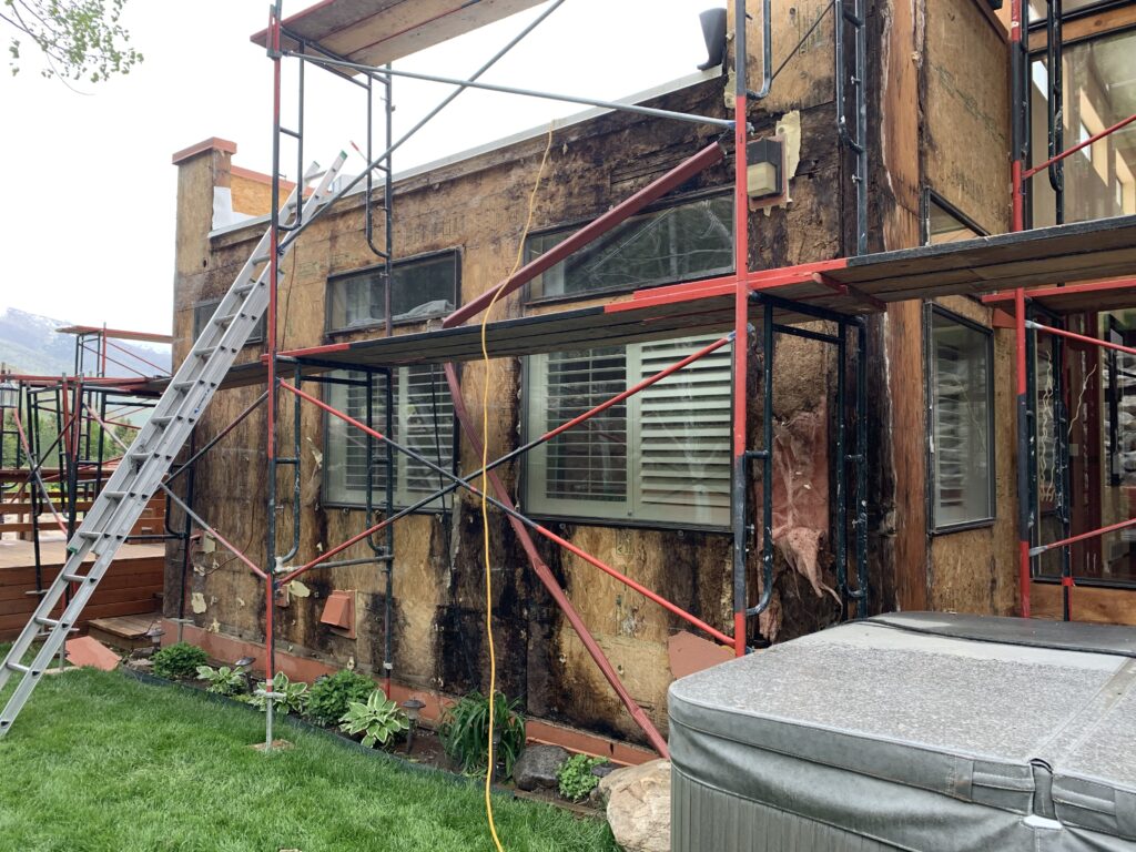 Intermediate stage of a massive structural remediation in Ogden, UT. Red scaffolding frames a home's exposed, severely water-damaged plywood sheathing. Piling of water-damaged sheathing and demoed building materials lies on the ground, illustrating a total structural fail.