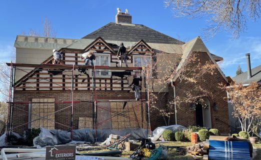 Close-up photo of roof demolition in progress, showing workers removing old dark asphalt shingles to expose the original roof deck.