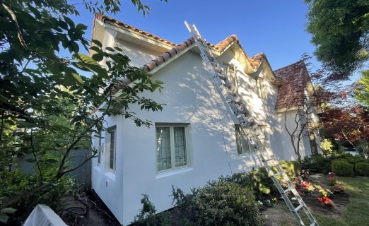An intermediate view of the house showing the new EIFS stucco system application in progress, with thick insulation boards visibly attached to the ground-floor brick walls to build the surface out.